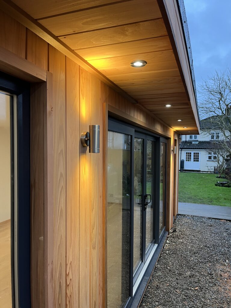 This image is an example of one of our garden rooms at dusk showcasing the brushed chrome downlights in the rich cedar canopy overhang and the up/down lights either side of the anthracite grey UPVC sliding patio door set. The lighting really brings out the beautiful warm tones of our cedar cladding. Highlighting also the expert carpentry shown by our team - all handcrafted on site by one small expert team of careful craftsmen.