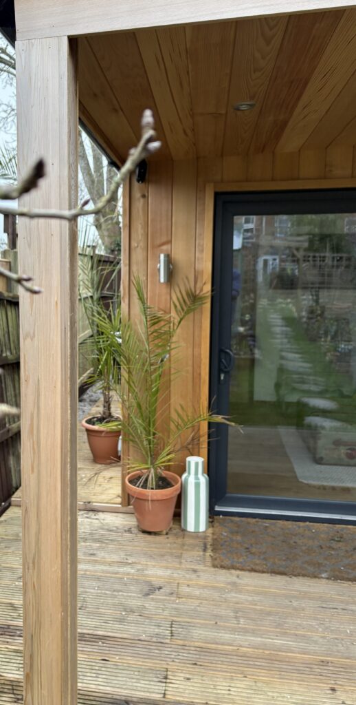 Showing side and front decking protected by a very deep cedar overhang with UPVC sliding doors opening into a sitting room. You can see brushed chrome up/down lights and downlights in the cedar canopy overhead. Potted plants soften the area.