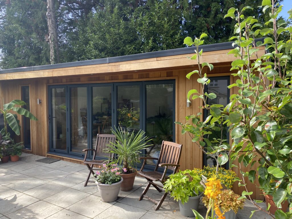 You can see a bespoke cedar clad garden room with large five pane bifold doors in anthracite grey with a full height window to each side of the bifold doors also in anthracite grey. The garden room is finished with a black rubber roof. There is a paved patio area to the front with wooden seating and lots of exotic plants in pots.