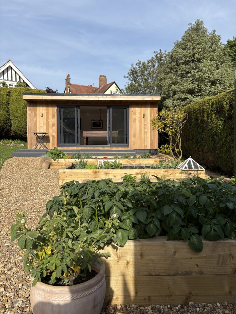 The raised vegetable beds in front of this garden room with four pane sliding patio door set in anthracite grey UPVC and silver up/down lights either side of the patio door set and black decking leading down to the garden area really make it feel part of the whole garden.