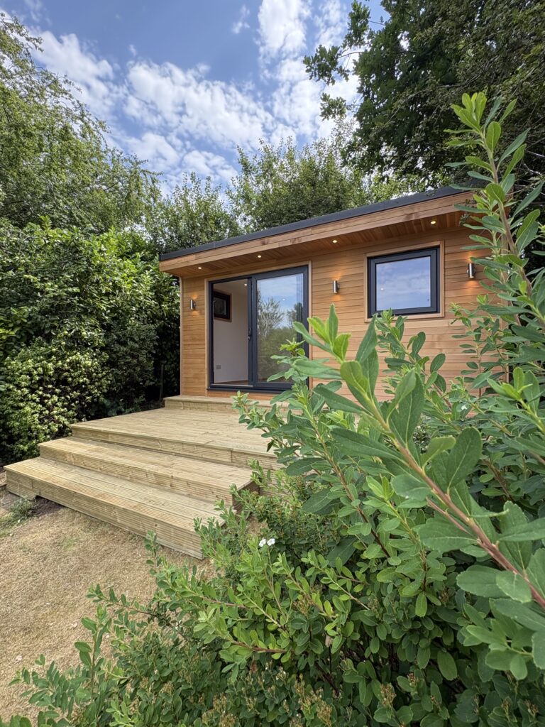 A cedar clad garden room with a square grey UPVC window and large grey sliding patio doors. The doors are open showing a white internal room. There are four downlights in the cedar clad canopy overhang. There are up/down lights next to the doors and the window. There is decking in several layers in front of the garden room and green shrubs surrounding it.