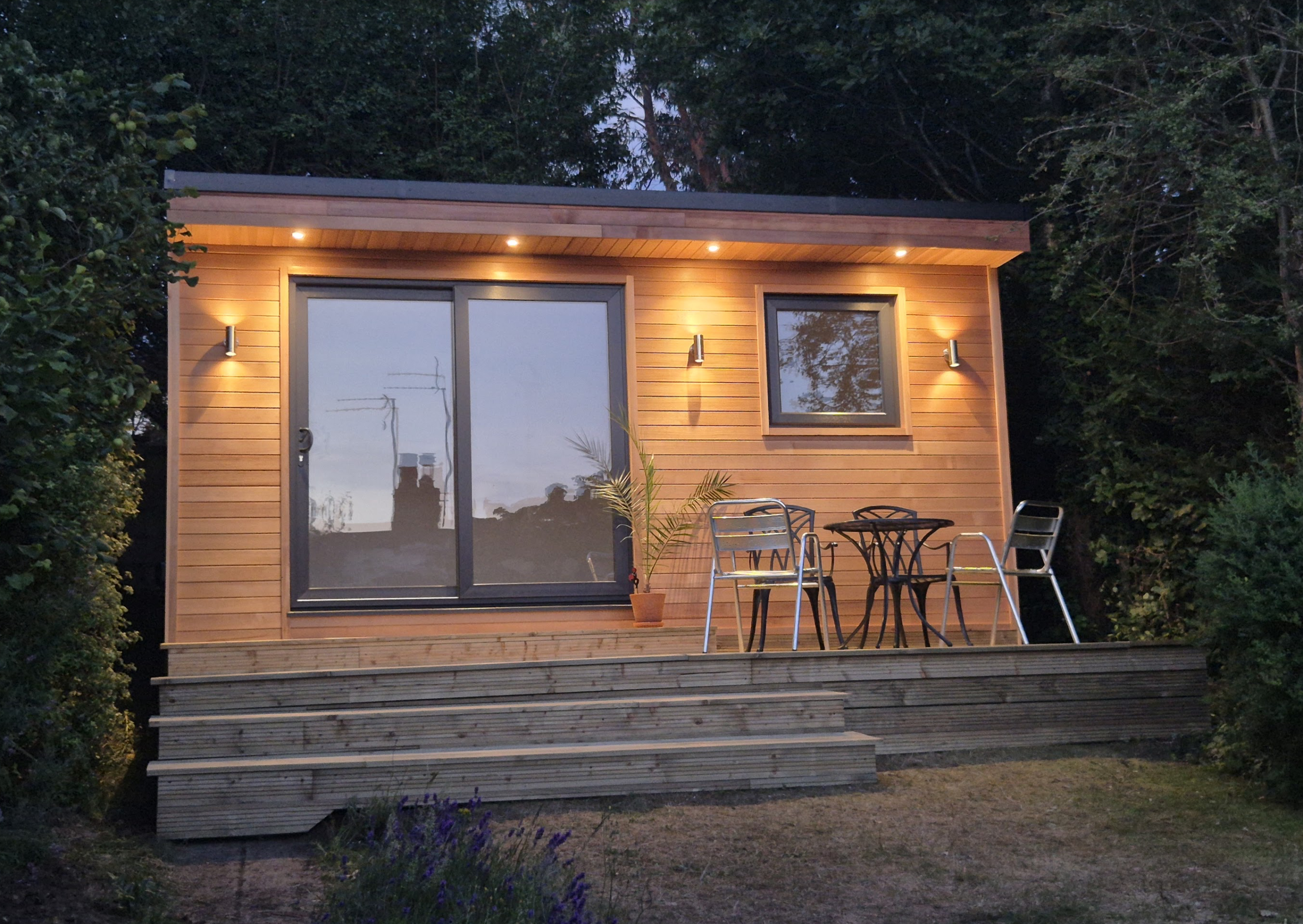 A cedar clad garden room with a square grey UPVC window and large grey sliding patio doors. There are four downlights in the cedar clad canopy overhang. There are up/down lights next to the doors and the window. There is decking in several layers in front of the garden room and green shrubs surrounding it.