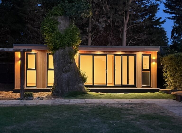 Image showing a cedar clad garden room lit up at night with a large tree in front. There is a siding patio door set and three floor to ceiling windows. The garden building has up/down lights at each end.